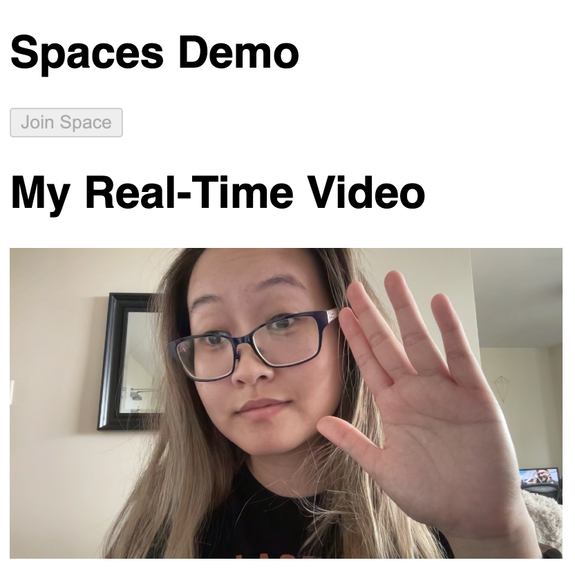 Capture input for real-time video that depicts Thuy, a young adult Asian woman, with long light brown hair and rectangular glasses, with her hand up as if about to wave to the viewer